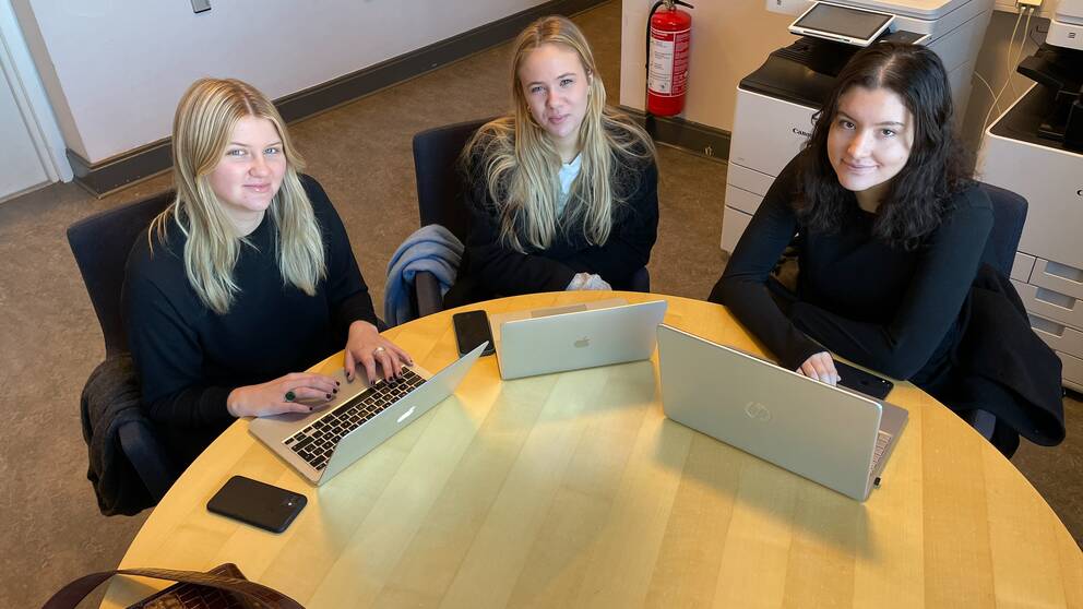 Tre kvinnliga studenter sitter runt ett bord med datorer framför sig. 