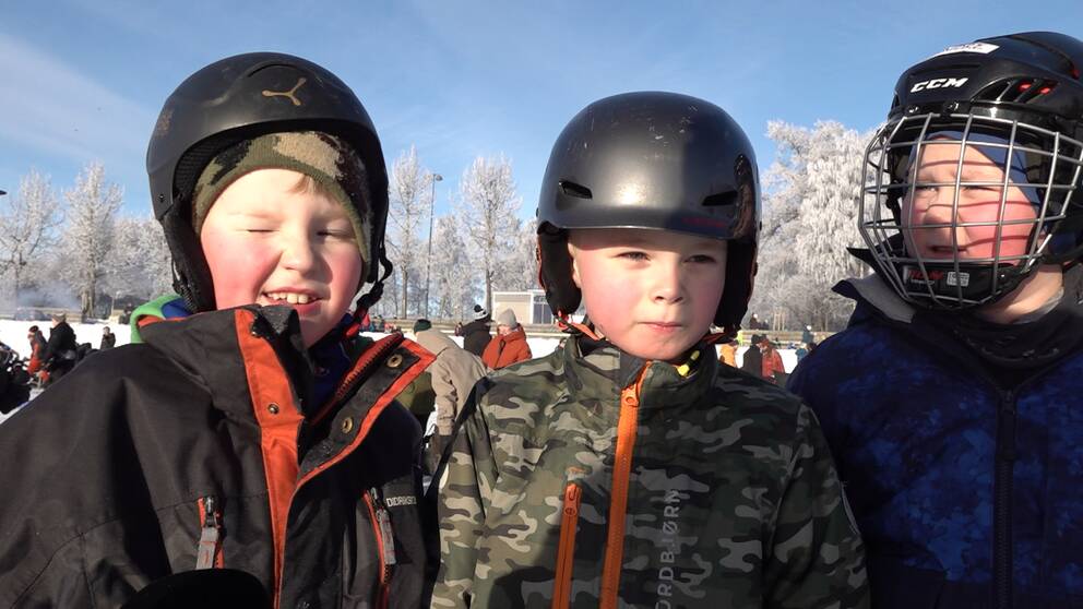 Tre pojkar med skridskohjälm kisar i solen utomhus.