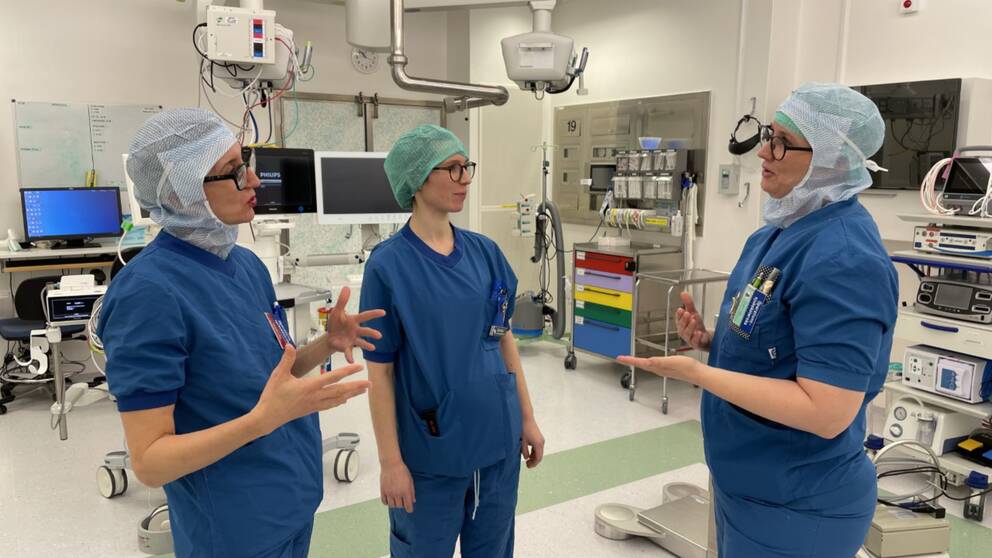 Tre sjuksköterskor i arbetskläder pratar med varandra och gestikulerar.
