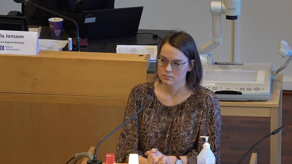 En kvinna i brun page och brun paisleymönsterad blus står bakom en mikrofon och ser ledsen ut.