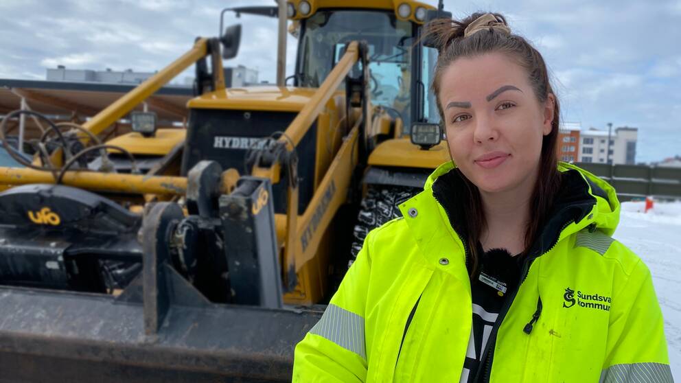 Kvinna i gul reflexjacka står framför en gul traktor.