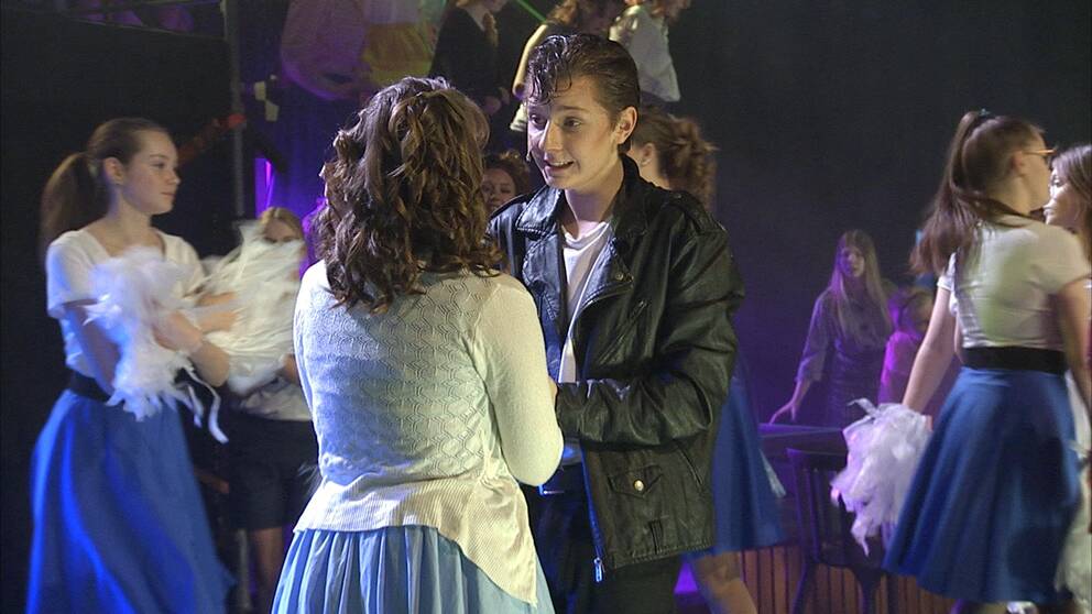 Två ungdomar står med händerna mot varandra, de spelar Sandy och Danny i Grease. Flera andra ungdomar med 50-talskjolar är runt omkring.