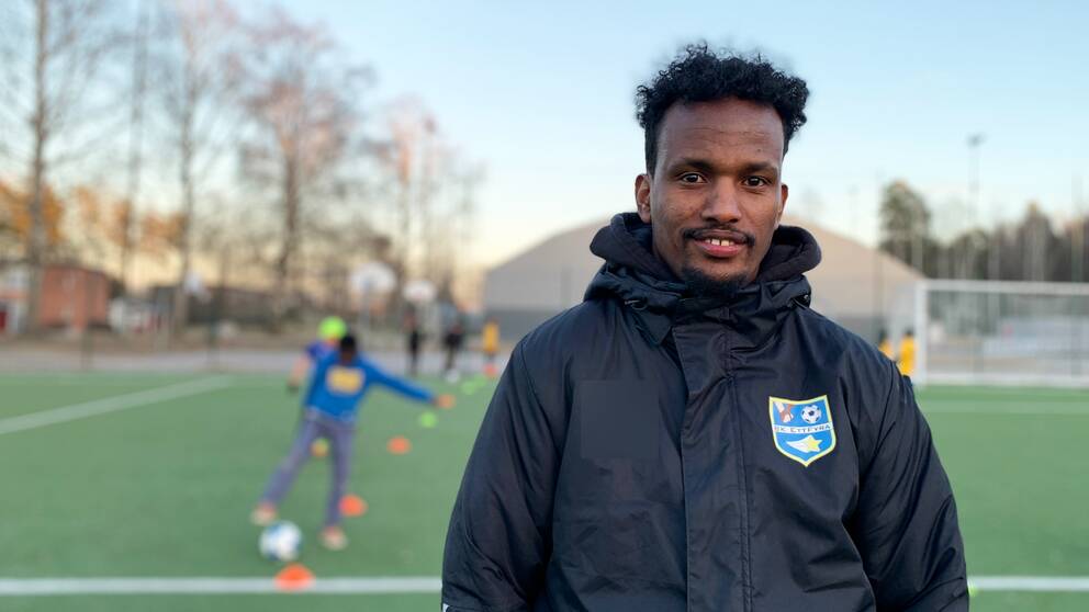 Fotbollstränaren och grundaren av fotbollsklubben BK Ett Fyra, Daud Awees, står på en fotbollsplan i klubbjackan medan det pågår en träning bakom honom.