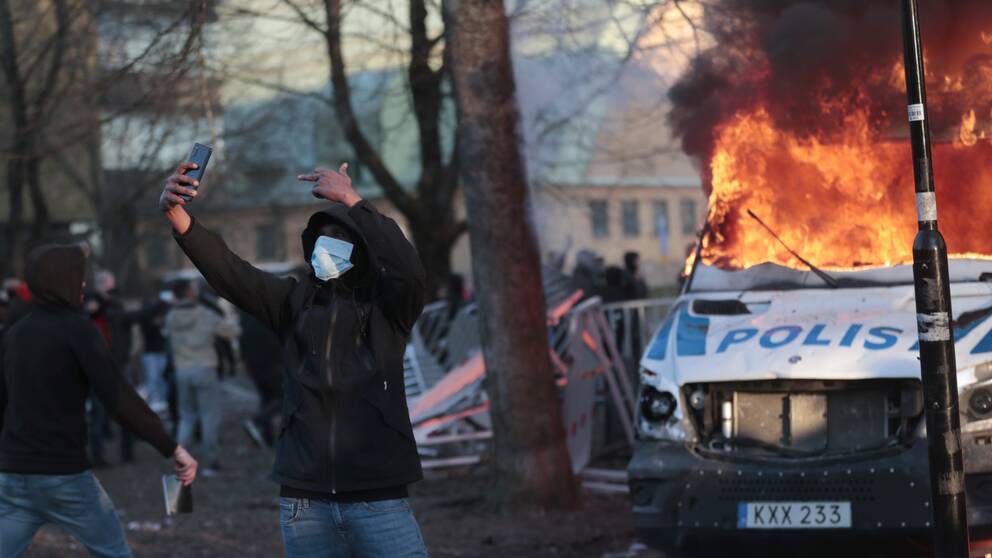 En man i maskering tar en selfie på sig själv och en brinnande polisbil i bakgrunden under påskupploppen i Örebro.