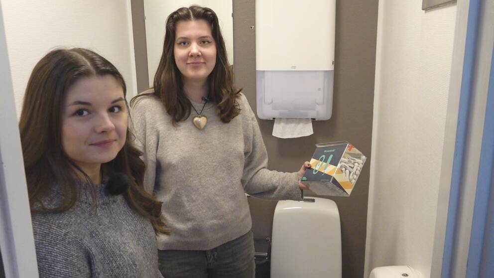 Två tjejer står inne på en toalett, den ena tjejen håller i en genomskinlig låda med tamponger