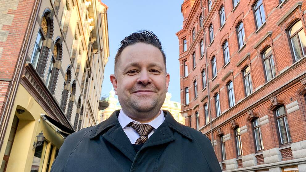 En bild på en Johannes Hulter som ser glad ut, och står framför två gamla tegelbyggnader i Göteborg. 