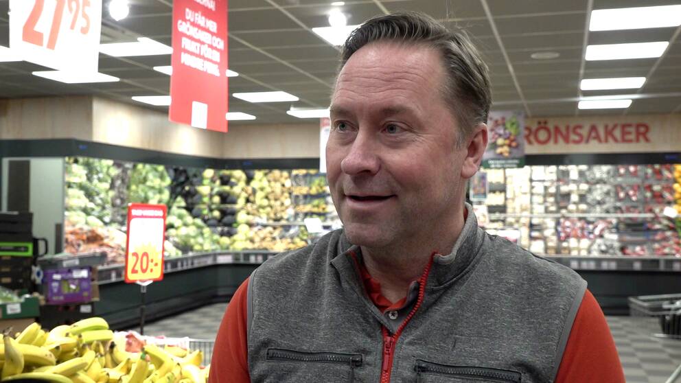 Handlaren Tomas Aronsson, en man i medelåldern, står iklädd arbetskläder framför grönsaksavdelningen i butiken han driver i Sundsvall.