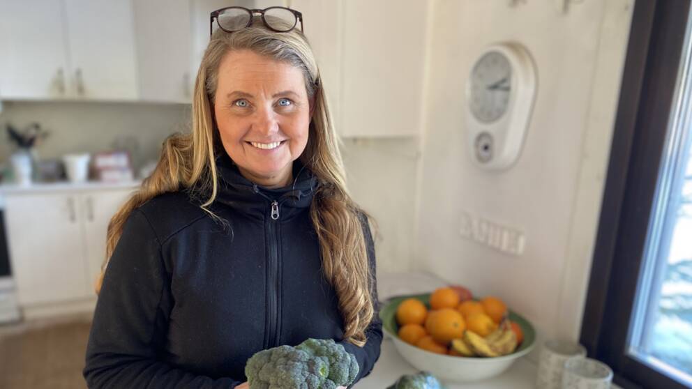 Det är en porträttbild av Malin Lindgren Sundström, som står i sitt kök och håller i en broccoli. Hon tittar och ler mot kameran.