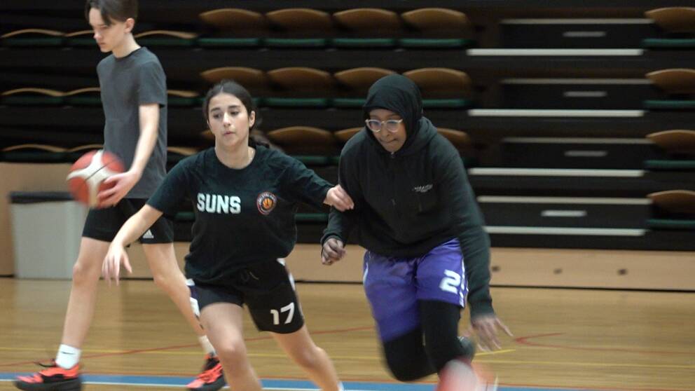 Två unga tjejer spelar basket på i en idrottshall.