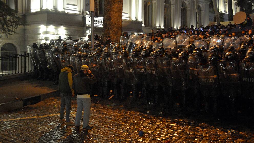 Two protesters stand in front of a sea of ​​policemen wearing protective gear. 