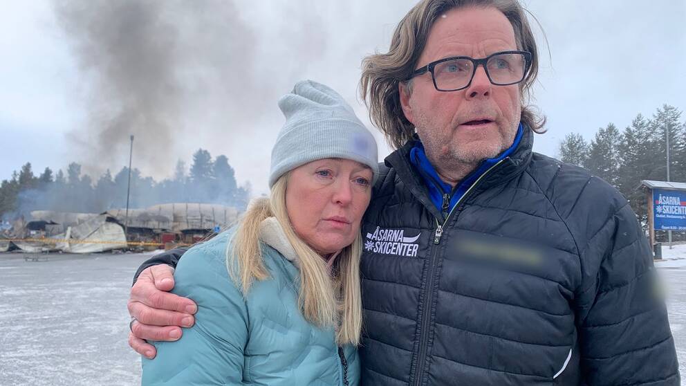 En blond kvinna med ljusblå jacka och grå mössa hålls om av en man med halvlångt hår och glasögon klädd i svart jacka. Bakom dem en en nedbrunnen byggnad.