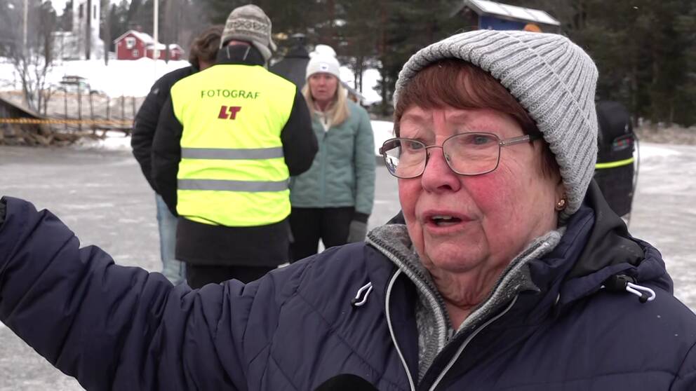 En kvinna i svart jacka och grå mössa står bredvid en brandplats och pekar. I bakgrunden ser man människor.