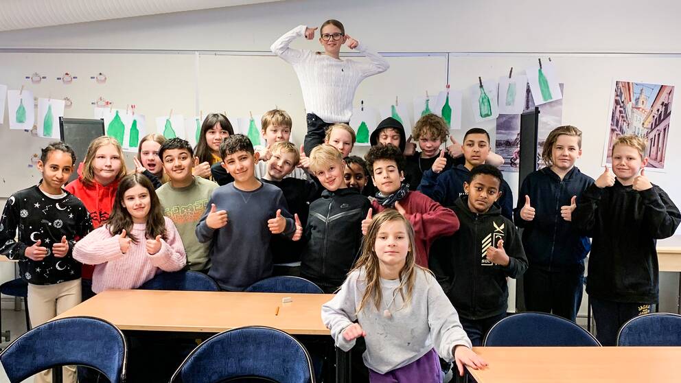 En bild på cirka 20 barn i ett klassrum, de ser glada ut och gör tummen upp. 