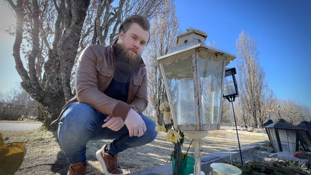 Bild på man med skägg och jacka som sitter på huk bredvid en grav. Mannen heter Kim Andersson och förlorade sin flickvän som valde att avsluta sitt liv för snart fyra år sedan. Han kritiserar avsaknaden av suicidprevention i Örebro läns kommuner i videoklippet.