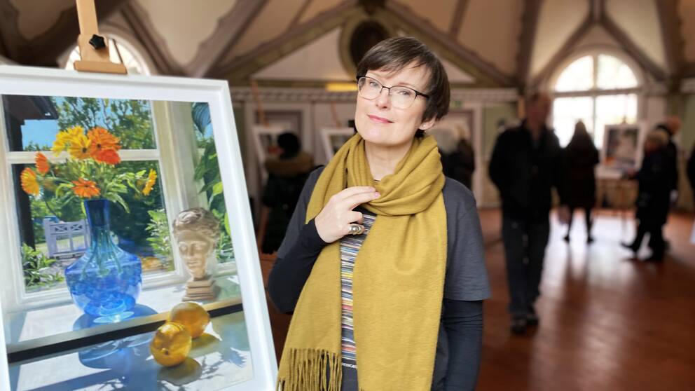 Bild på en målning och besökare i en utställningslokal. Bredvid målningen står en kvinna, som heter CajsaStina Åkerström och är konstnär.