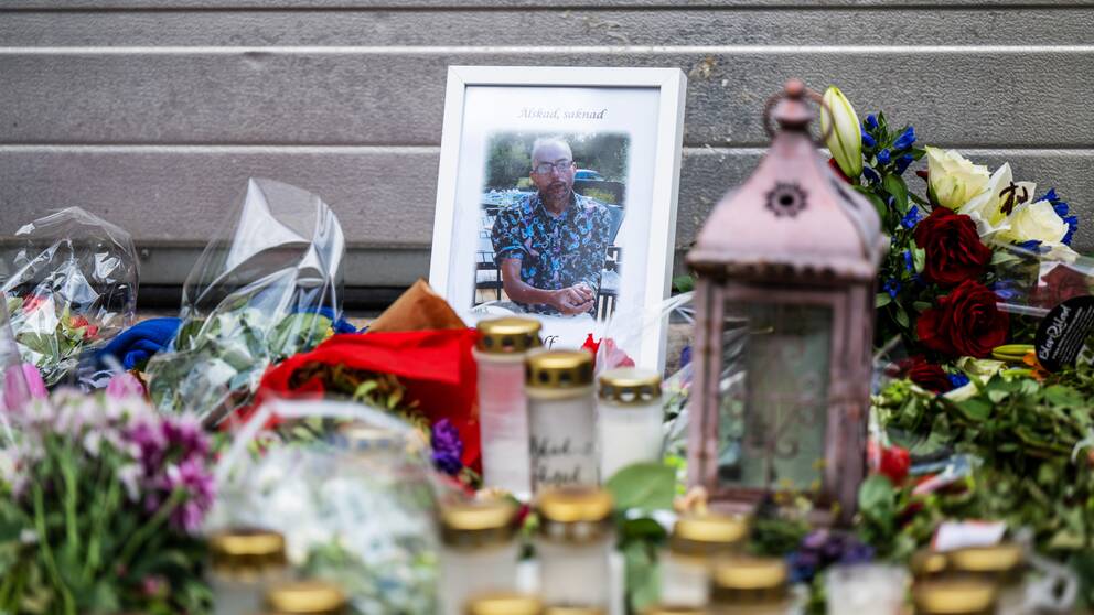 Ett fotografi av den mördade mannen Ulf Sandberg vid mordplatsen, med texten ”Älskad, saknad” och en stor mängd blommor och ljus.