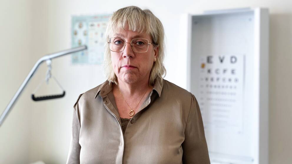 Ingmarie Kasche står i sitt arbetsrum. I bakgrunden syns en brits och en karta med bokstäver i olika storlekar som används vid synundersökningar.