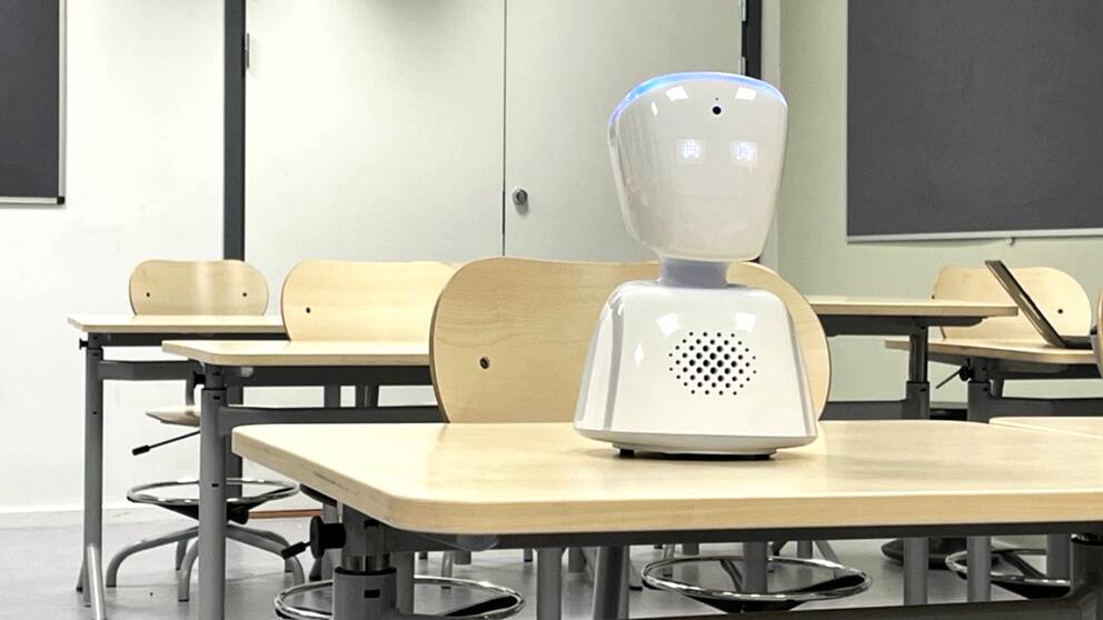 En vit oformlig liten robot står i ett klassrum med glada ögon.