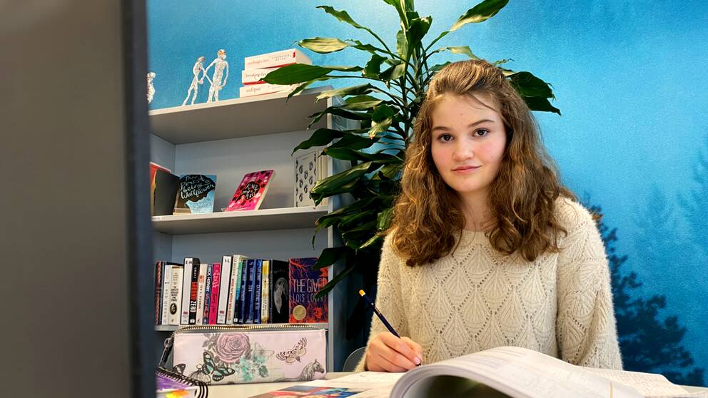 13-åringen Emilia Kenaga sitter vid ett arbetsbord med en stor grön växt bakom sig och har skolböcker framför sig och en penna i handen.