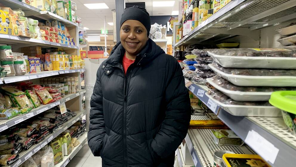 Fatime Noor, står mellan två hyllor mat i en matbutik, är mörkhyad, har svart huvudbonad och jacka, röd tröja, hon ler mot kameran.