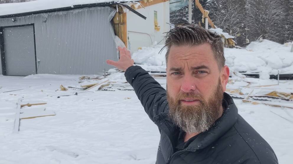 Vd:n Antony Viklund, står inför det raserade taket och pekar mot den del av byggnaden som består. Viklund har en svart jacka och skägg och står i ett snöigt landskap i Robertsfors kommun.