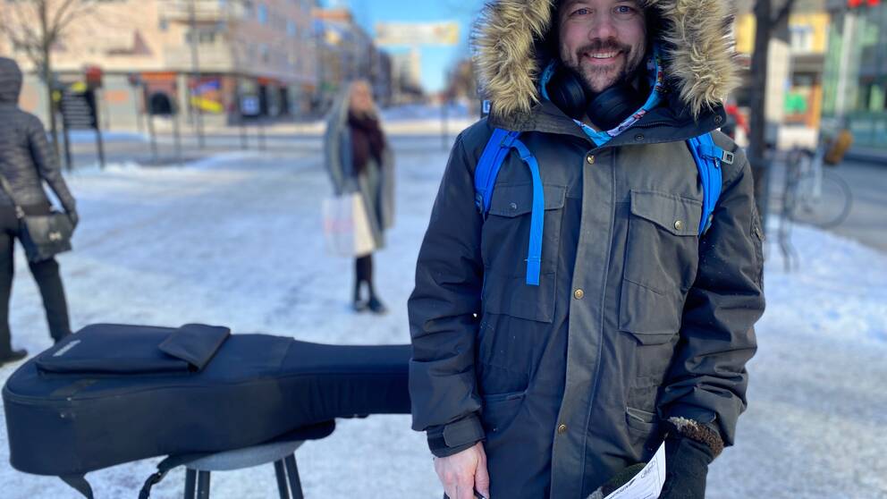 Anton Boström står iklädd vinterjacka med pälskrage och ler. Han har mörkt kort hår och skägg. Snett bakom honom längs gågatan i Luleå ligger gitarren på en pall.