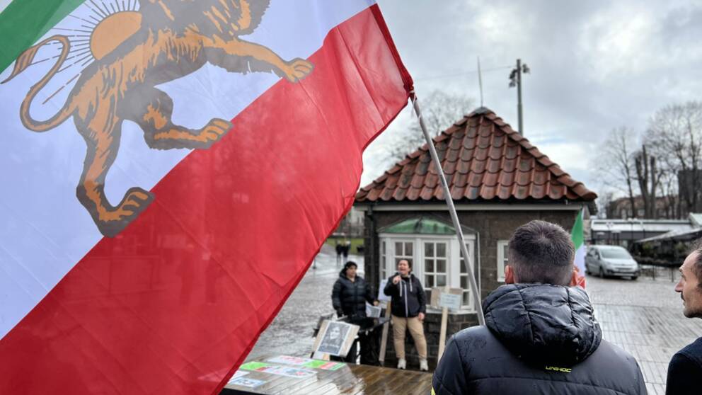 En stor flagga från Iran täcker vänstra sidan av bilden samtidigt som man ser en rygg på en ung kille som håller i flaggan och kollar framåt.