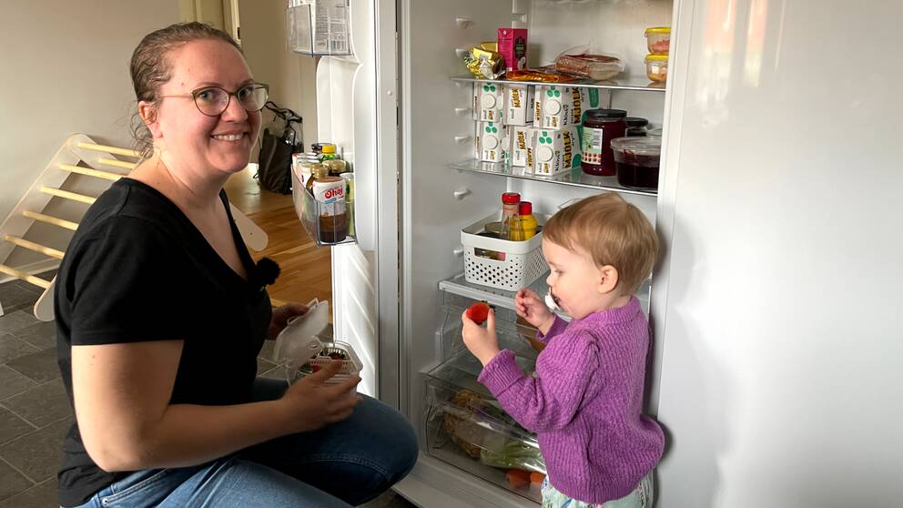 Klara Twete sitter på huk framför kylen och ger sin dotter en jordgubbe.