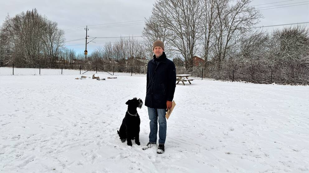 Sam Graflund Wallentin, en man i jacka och mössa, står ute i snön på en hundlekplats tillsammans med sin hund.