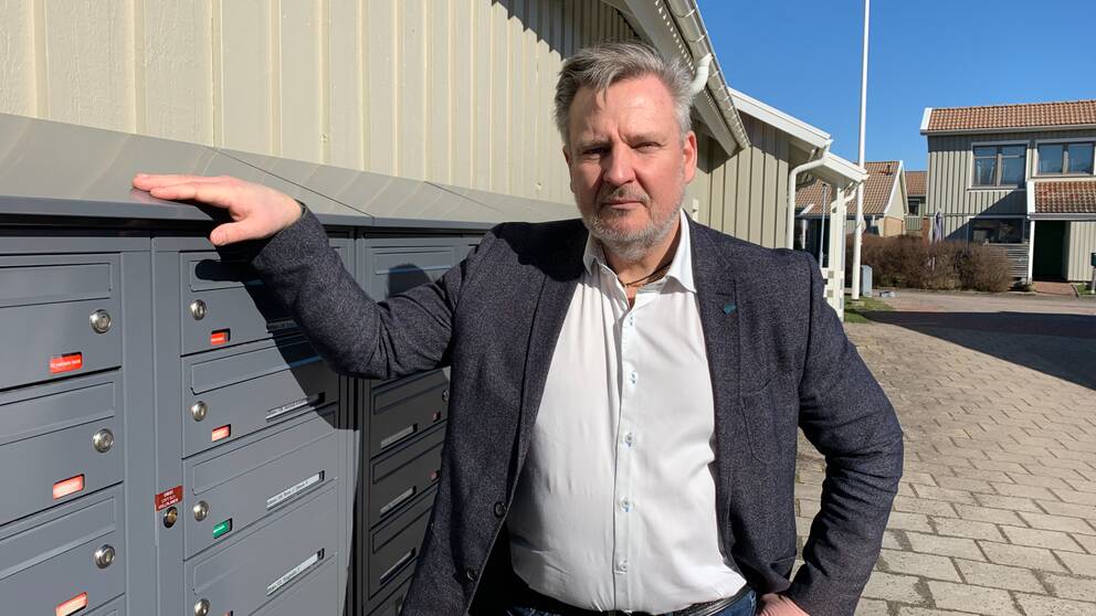 En man står med ena handen på en postboxanordning. Bakom honom finns rashus.