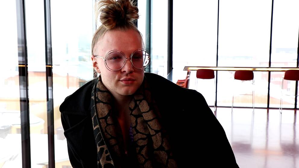 En ung man med håret knutet ovanpå huvudet sitter framför en glasvägg.