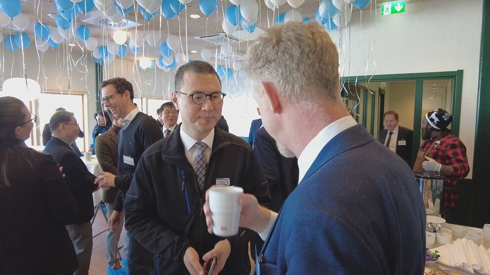 Vita och blå ballonger hänger taket. Flera personer står och pratar med varandra och ser glada ut. Donjings platschef Yun Lee lyssnar på en man som håller i en kaffekopp.