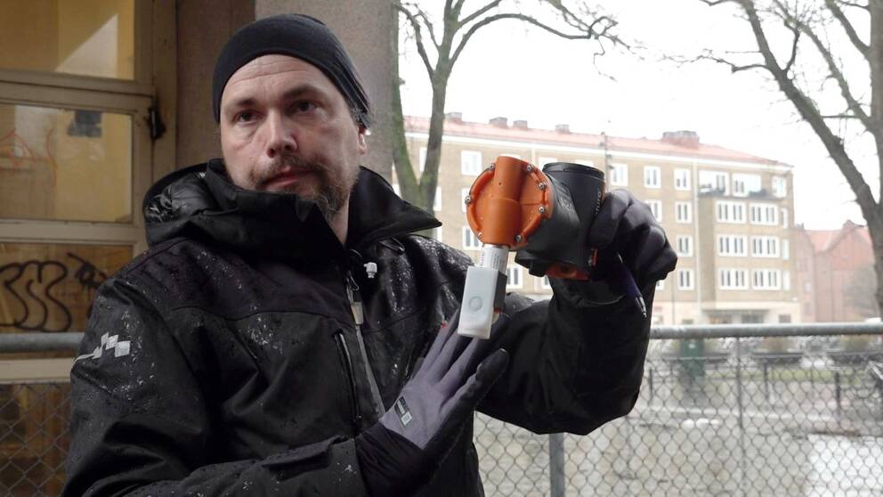Skadedjurstekniker Anders Israelsson visar upp en råttfälla. Han står utomhus framför en byggnad och ett staket.