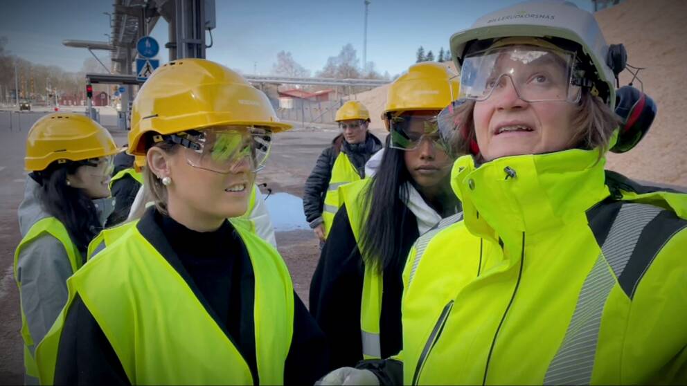 En kvinnlig ingenjör står omringad av unga tjejer i gula hjälmar och reflexvästar.