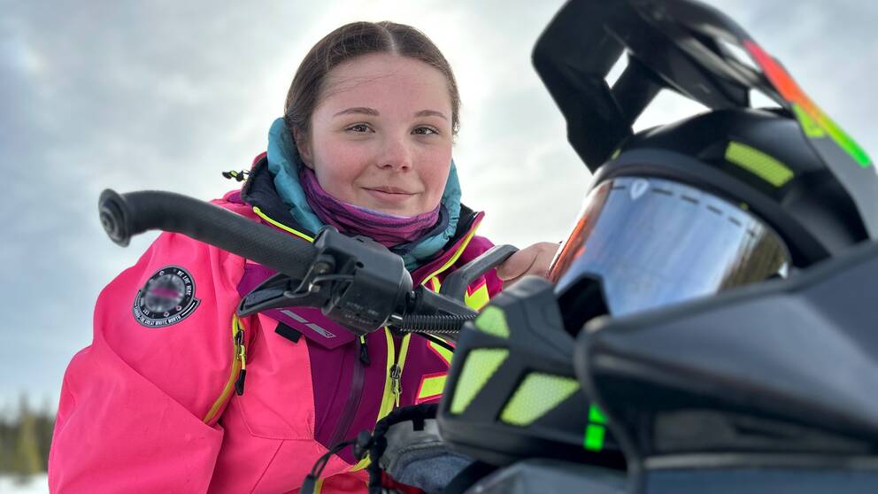 19-åriga Irma Åkerblom, en mörkhårig tjej iklädd rosa skoterkläder sitter på sin skoter i Storuman med hjälmen på styret och ler in i karmeran.