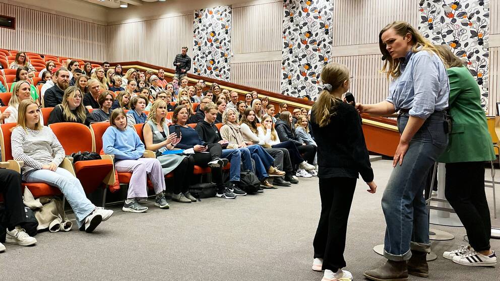 Elever sitter som publik i röda stolar bakom en scen där en kvinna med en blå skjorta intervjuar ett barn.