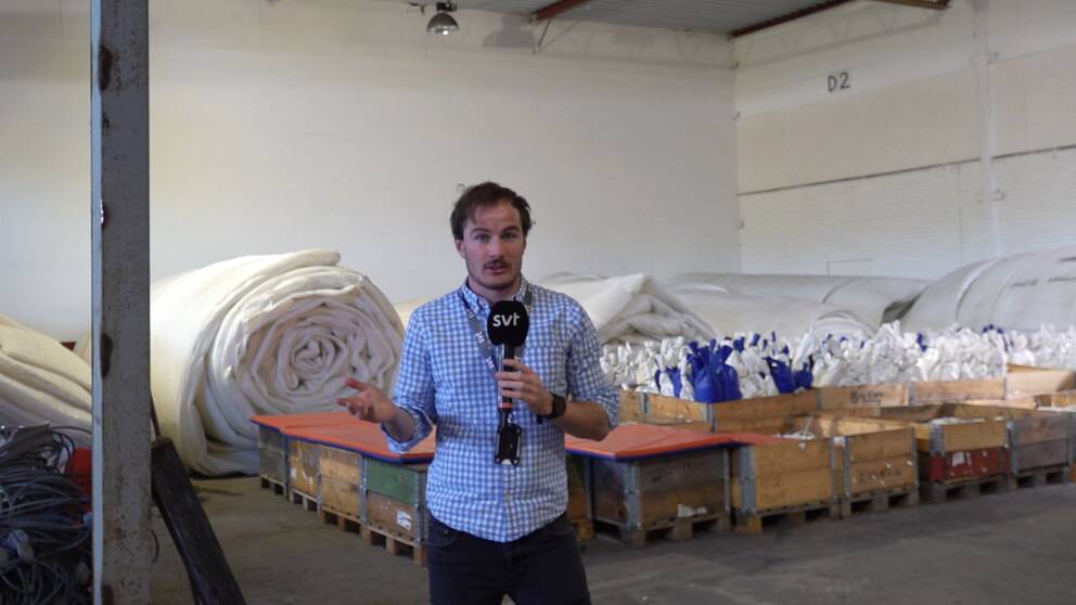 Reportern står framför ihopprullad folie och sandsäckar i ett stort lagerlokal.