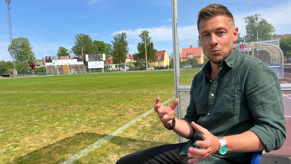 En man i grön skjorta sitter i avbytarbåset vid en fotbollsplan.
