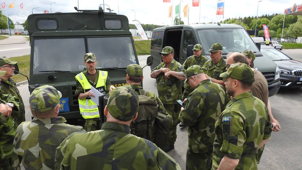 Hemvärnssoldater står samlade kring ett militärfordon vid en bensinmack. En man iklädd reflexväst med texten ”militär insatschef” håller en dragning och de övriga lyssnar  uppmärksamt.