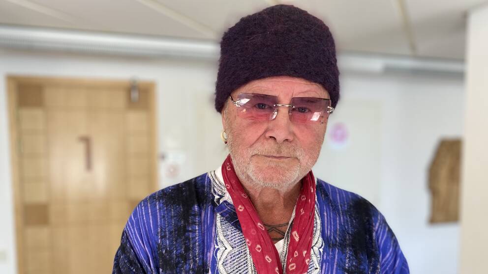 På bilden står Roger Pontare. Han har glasögon och en mörk mössa på sig. Han tittar in i kameran och har en blå/lila tröja och en röd halsduk.