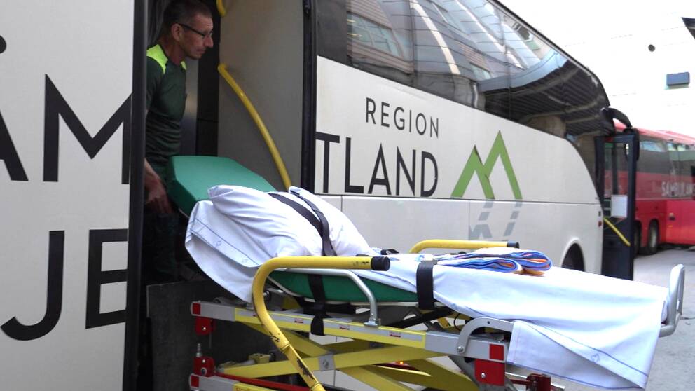 En ambulanssjukvårdare står i den bakre dörren av en vit buss med svart text. Han håller i en sjukhusbår och tittar ut åt sidan, bort från kameran.