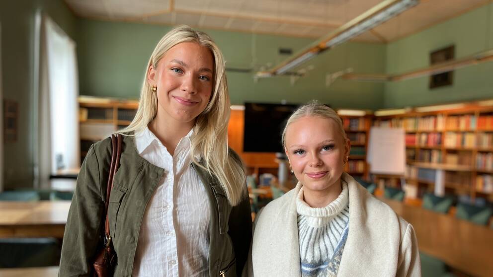 Tyra Nordvall och Liv Sandell Jonsson, två tjejer med blont hår står i en skollokal med bokhyllor i bakgrunden och tittar in i kameran.