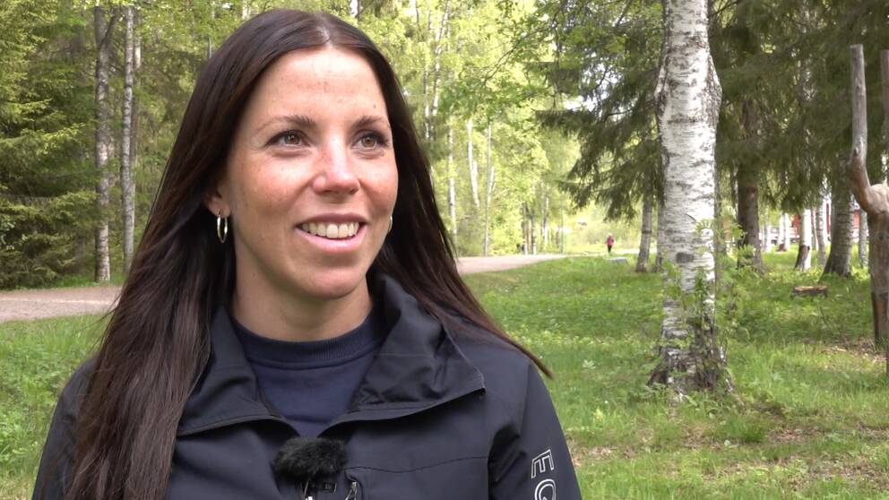 På bilden är Charlotte Kalla. Hon ler så att tänderna ses, hon har runda örhängen på sig och brunt hår. Hon tittar inte in i kameran och hon står vid gräs och träd.