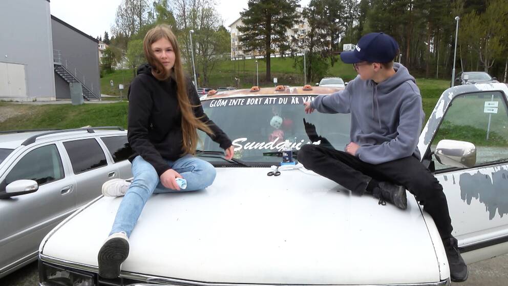 Två ungdomar – en tjej och en kille – sitter på en vit motorhuv. Killen pekar på dekalen i ovankanten på vindrutan, det står ”Kom ihåg att du är värdefull”.