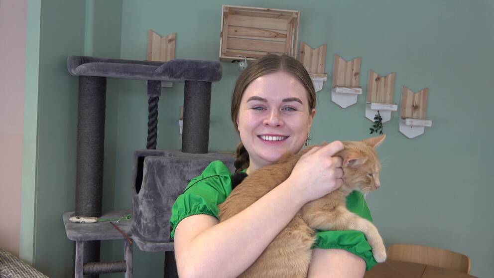 En kvinna ler och tittar rakt in i kameran. I famnen håller hon en röd katt som spinner. Hon har en grön tröja och i bakgrunden ses ett klätterträd för katter.
