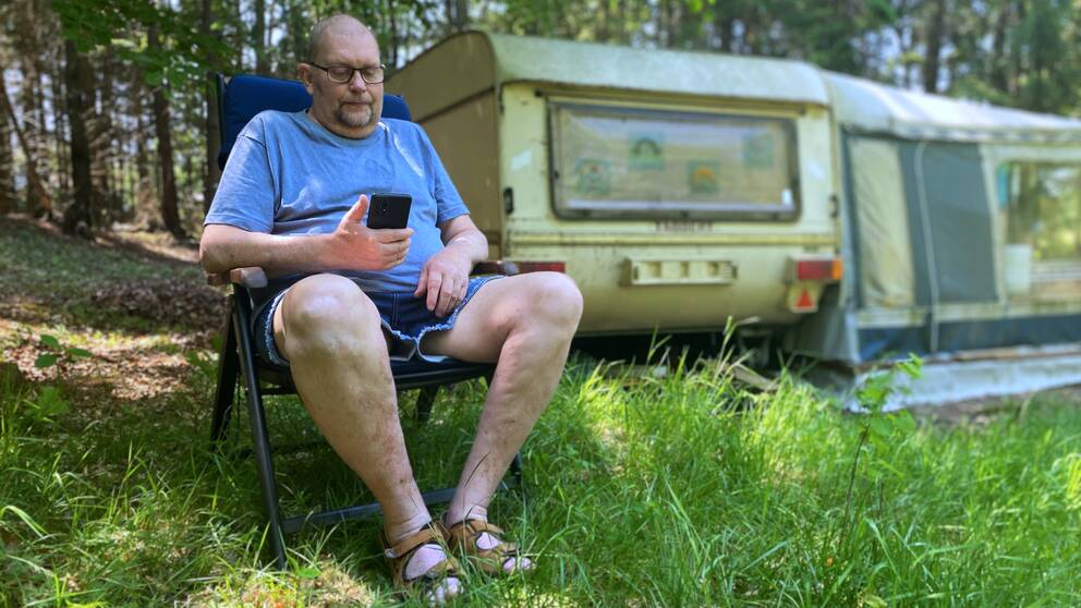 Bill Lundberg sitter i solstol utanför sin husbil och tittar i sin mobil.