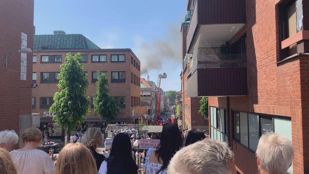Folksamling står och kollar på branden i ett lägenhetshus och studentflak passerar i bakgrunden.