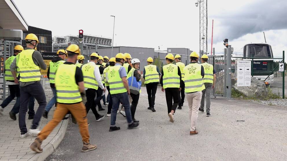 Folksamling på ca 20 personer i varselvästar och gula hjälmar går in genom grinden till en industrianläggning som syns i bakgrunden.