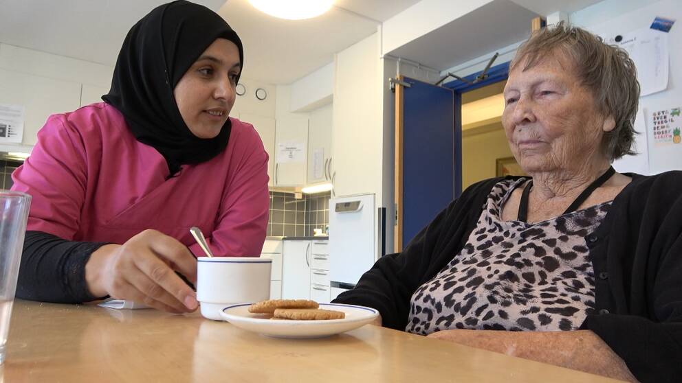 En kvinna i sjal och rosa tröja serverar en äldre kvinna kaffe och kakor vid ett bord