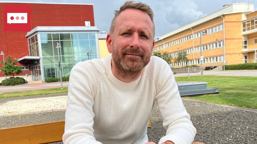 En man i vit tröja sitter på en bänk utomhus och tittar in mot kameran med ett litet leende.
